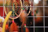 A close-up view of colorful dreamcatchers hung against a metal grid. The design features brightly colored nets and feathers, with beads adding decorative elements. The background is softly blurred, highlighting the vibrant details of the dreamcatchers.