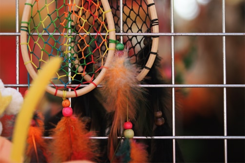 A close-up view of colorful dreamcatchers hung against a metal grid. The design features brightly colored nets and feathers, with beads adding decorative elements. The background is softly blurred, highlighting the vibrant details of the dreamcatchers.