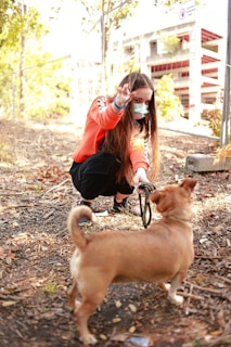 A person wearing an orange sweater and a face mask is crouching down in a wooded area, interacting with a small brown dog. The person is holding the dog's leash while making a peace sign gesture with one hand. Sunlight filters through the trees, creating a warm atmosphere.