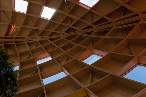 An intricate wooden architectural structure featuring a grid-like pattern of beams and panels against a sky-blue background. The design creates a geometric and artistic display, with some leafy green plants visible on the left side.