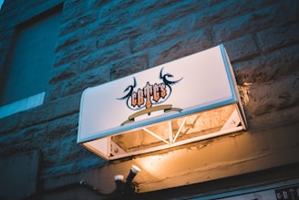 An outdoor sign with ornate text and artistic flourishes, mounted on a stone wall. The sign is illuminated, casting a subtle glow on the surrounding bricks. The background features a textured stone surface, adding a rustic feel.