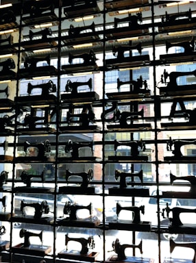 Rows of vintage sewing machines are displayed on shelves in front of a window, creating a patterned silhouette. The background shows an urban street scene with buildings and parked cars.