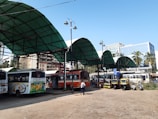 A bus station with several buses parked under large green canopy roofs. The area is bustling with activity, featuring a few autorickshaws and scooters nearby. People are seen walking around, engaging in their daily commute. In the background, there are tall buildings and palm trees, indicating an urban environment. The sky is clear, suggesting pleasant weather.