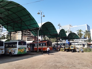 A bus station with several buses parked under large green canopy roofs. The area is bustling with activity, featuring a few autorickshaws and scooters nearby. People are seen walking around, engaging in their daily commute. In the background, there are tall buildings and palm trees, indicating an urban environment. The sky is clear, suggesting pleasant weather.