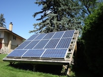 A large, ground-mounted solar panel array is positioned in a grassy area with trees and a house nearby. The panels are tilted to capture sunlight efficiently.