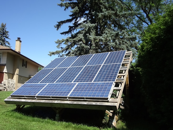 A large, ground-mounted solar panel array is positioned in a grassy area with trees and a house nearby. The panels are tilted to capture sunlight efficiently.