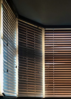 Vertical blinds partially open, allowing light to filter through, creating a pattern of light and shadow on the wooden slats. A mix of warm and cool tones is visible, with contrasting shades on each side influenced by the angle of the light.