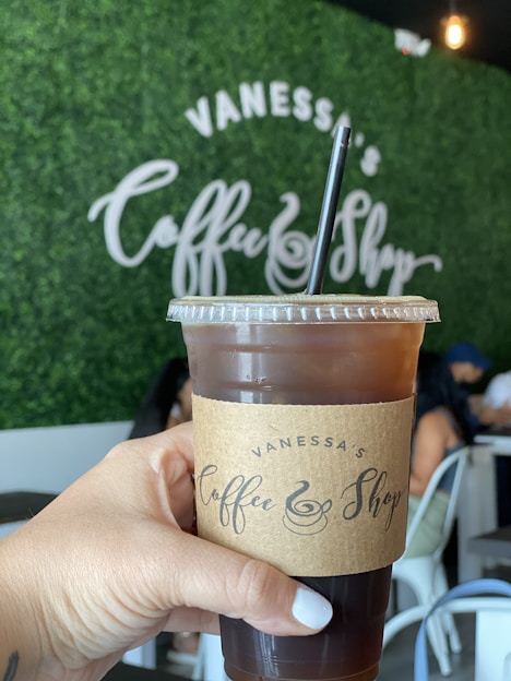 A hand holds an iced coffee in a clear cup with a brown cardboard sleeve labeled 'Vanessa's Coffee Shop'. The background features a green leafy wall with the shop's name written in large white letters.