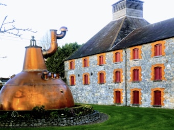 A large copper distillation apparatus is prominently placed in front of a historic stone building with red-framed windows and a steep roof. The well-maintained lawn adds a touch of greenery to the scene, and the sky is clear, indicating pleasant weather.