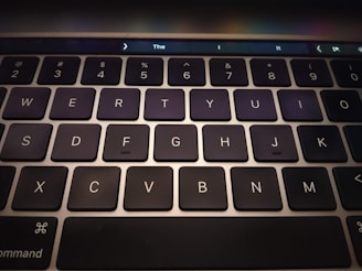 A close-up view of a computer keyboard featuring black keys with white letters and numbers. The keys are arranged in a standard QWERTY layout, and a touch bar is visible at the top of the keyboard, displaying some icons and text.
