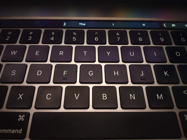 A close-up view of a computer keyboard featuring black keys with white letters and numbers. The keys are arranged in a standard QWERTY layout, and a touch bar is visible at the top of the keyboard, displaying some icons and text.