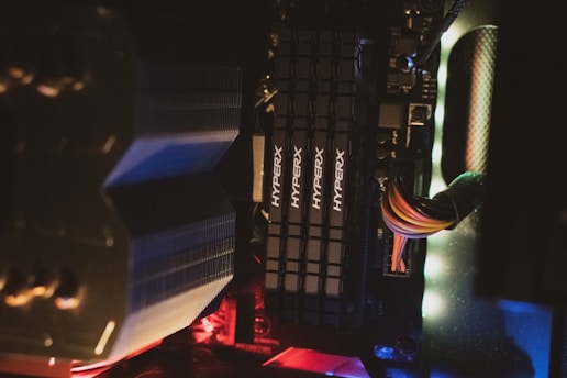 Close-up view of computer components including multiple RAM sticks labeled HyperX, an array of colored cables, and a metallic cooling unit, all housed within a computer case illuminated by lights.