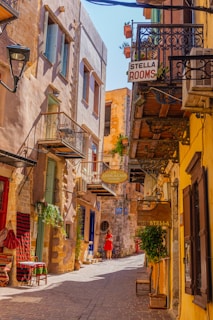 A narrow, picturesque street with charming stone and stucco buildings on either side. Balconies with ornate iron railings and potted plants add to the quaint atmosphere. Signs for accommodations hang from the buildings, and a person in red is walking down the sunlit street. The ground is covered with cobblestones, enhancing the historic and cozy feel.