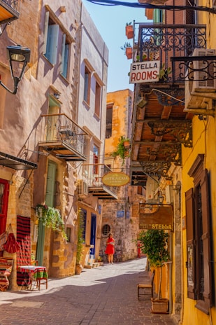 A narrow, picturesque street with charming stone and stucco buildings on either side. Balconies with ornate iron railings and potted plants add to the quaint atmosphere. Signs for accommodations hang from the buildings, and a person in red is walking down the sunlit street. The ground is covered with cobblestones, enhancing the historic and cozy feel.