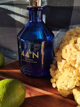 A blue glass bottle labeled '44°N Gin Imagined and Distilled in Grasse' is placed on a wooden surface. It is accompanied by fresh green limes and a cluster of small yellow flowers. The scene captures sunlight, casting soft shadows and adding warmth to the composition.