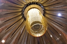 An ornate chandelier with intricate metallic patterns extends from the ceiling, featuring a central glass component illuminated with warm light. The design radiates outward with a fan-like arrangement, showcasing detailed craftsmanship and a luxurious feel.