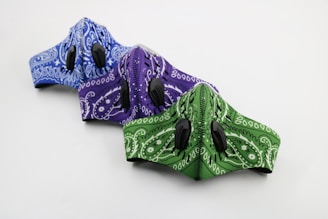 Three patterned face masks in blue, purple, and green are aligned in a row. Each mask features a paisley design and has two black air valves.