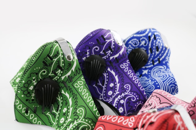A collection of cloth masks featuring vibrant paisley patterns in different colors, including green, purple, blue, and red. Each mask has a black valve and a metal nose clip for a better fit. The patterns and bright colors create an eye-catching display.