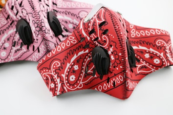 Two intricately designed face masks featuring paisley patterns. One mask is primarily red with bold white and black designs, and the other is pink with similar patterns. Both masks have black breathing valves.