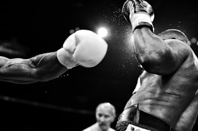A dynamic black and white photograph captures an intense moment in a boxing match. One boxer delivers a punch to their opponent, causing droplets of sweat to fly. The muscular form of the boxers is clearly visible, highlighting their physical prowess.