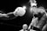 A dynamic black and white photograph captures an intense moment in a boxing match. One boxer delivers a punch to their opponent, causing droplets of sweat to fly. The muscular form of the boxers is clearly visible, highlighting their physical prowess.