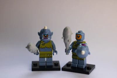 Two blue toy figurines with cyclopean features and primitive outfits stand side by side on black bases. Each holds a club, and they exhibit cartoonish features like single large eyes and exaggerated expressions.