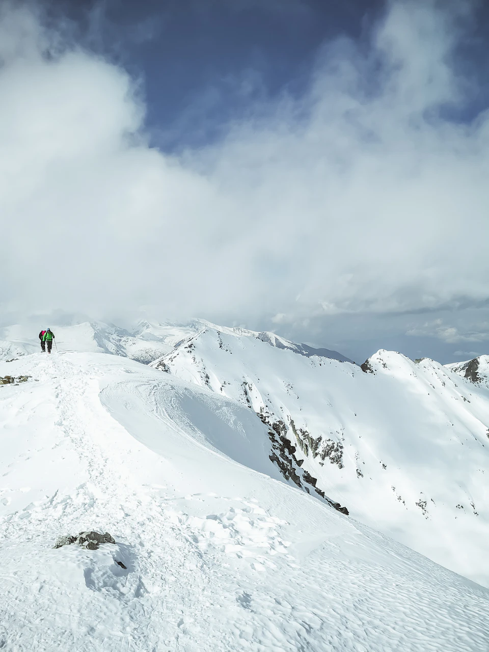 The image depicts a snowy mountain peak with two hikers making their way up, one closer to the camera and another further away on the left side of the frame. The sky above them is filled with clouds, creating a dramatic backdrop for the scene. The snow-covered mountains in the background are covered in white, providing a stark contrast against the clear blue sky.

The hikers appear to be moving towards the right side of the image, their figures slightly blurred due to the distance and motion. The overall composition suggests that they are on an outdoor adventure, exploring the beauty of nature at this snowy peak.