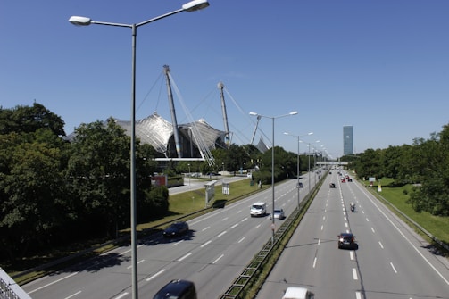 A wide highway with multiple lanes is flanked by a series of tall street lights. On the left side, a unique architectural structure with a tent-like design is visible, featuring large glass and steel elements. Trees and greenery line both sides of the road, while a few vehicles are spread out along the highway under a clear blue sky.