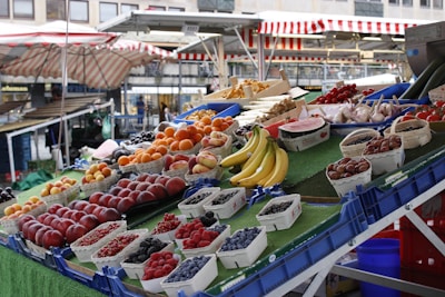 A vibrant market stall featuring an array of fresh fruits and vegetables. On display are bananas, berries, nectarines, apricots, cherries, watermelon, and garlic, all organized neatly in containers. The stall is set against a backdrop of striped market canopies and a bustling marketplace.