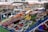 A vibrant market stall featuring an array of fresh fruits and vegetables. On display are bananas, berries, nectarines, apricots, cherries, watermelon, and garlic, all organized neatly in containers. The stall is set against a backdrop of striped market canopies and a bustling marketplace.