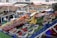 A vibrant market stall featuring an array of fresh fruits and vegetables. On display are bananas, berries, nectarines, apricots, cherries, watermelon, and garlic, all organized neatly in containers. The stall is set against a backdrop of striped market canopies and a bustling marketplace.