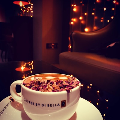 A cozy coffee shop environment featuring a cup of coffee topped with dried rose petals. The atmosphere is warm and inviting with dim lighting from string lights and reflections creating a soft glow. A comfortable chair adds to the relaxed setting.