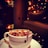 A cozy coffee shop environment featuring a cup of coffee topped with dried rose petals. The atmosphere is warm and inviting with dim lighting from string lights and reflections creating a soft glow. A comfortable chair adds to the relaxed setting.