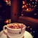 A cozy coffee shop environment featuring a cup of coffee topped with dried rose petals. The atmosphere is warm and inviting with dim lighting from string lights and reflections creating a soft glow. A comfortable chair adds to the relaxed setting.