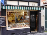 A quaint storefront with a green and white striped awning above a display window. The window shows various goods, possibly pastries or bread, with a person seen working inside. The door to the right is black and numbered 129. A chalkboard menu is displayed to the left of the shop.