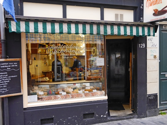 A quaint storefront with a green and white striped awning above a display window. The window shows various goods, possibly pastries or bread, with a person seen working inside. The door to the right is black and numbered 129. A chalkboard menu is displayed to the left of the shop.