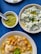 Three dishes are arranged on a blue table. One dish contains chickpeas and a poached egg in a broth garnished with a green herb paste. Another contains a creamy salad with cucumbers and herbs topped with olive oil. A small bowl has a green pesto-like sauce with a spoon.