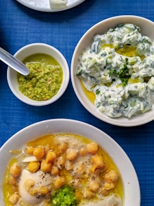 Three dishes are arranged on a blue table. One dish contains chickpeas and a poached egg in a broth garnished with a green herb paste. Another contains a creamy salad with cucumbers and herbs topped with olive oil. A small bowl has a green pesto-like sauce with a spoon.