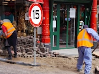 Two workers wearing orange safety vests are engaged in street maintenance or construction work. The area is surrounded by red pillars, and there is a number 15 speed limit sign nearby. A partially completed brick pavement and a shop entrance with glass doors can be seen in the background. Gold Buddha statues are displayed in the window of the shop.