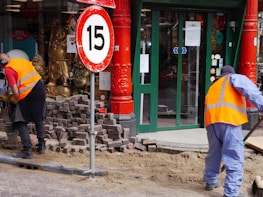 Two workers wearing orange safety vests are engaged in street maintenance or construction work. The area is surrounded by red pillars, and there is a number 15 speed limit sign nearby. A partially completed brick pavement and a shop entrance with glass doors can be seen in the background. Gold Buddha statues are displayed in the window of the shop.