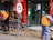 Two workers wearing orange safety vests are engaged in street maintenance or construction work. The area is surrounded by red pillars, and there is a number 15 speed limit sign nearby. A partially completed brick pavement and a shop entrance with glass doors can be seen in the background. Gold Buddha statues are displayed in the window of the shop.