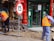 Two workers wearing orange safety vests are engaged in street maintenance or construction work. The area is surrounded by red pillars, and there is a number 15 speed limit sign nearby. A partially completed brick pavement and a shop entrance with glass doors can be seen in the background. Gold Buddha statues are displayed in the window of the shop.