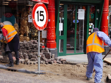Two workers wearing orange safety vests are engaged in street maintenance or construction work. The area is surrounded by red pillars, and there is a number 15 speed limit sign nearby. A partially completed brick pavement and a shop entrance with glass doors can be seen in the background. Gold Buddha statues are displayed in the window of the shop.