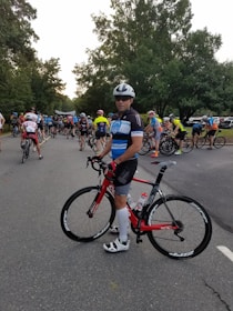 A group of cyclists gathered on a road, with a prominent cyclist in the foreground holding a red bicycle. The participants wear colorful cycling attire and helmets, preparing for what seems to be a race or an organized ride event. Tall trees line the road, creating a lush green backdrop.