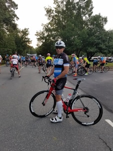 A group of cyclists gathered on a road, with a prominent cyclist in the foreground holding a red bicycle. The participants wear colorful cycling attire and helmets, preparing for what seems to be a race or an organized ride event. Tall trees line the road, creating a lush green backdrop.