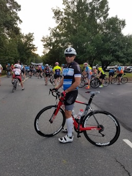 A group of cyclists gathered on a road, with a prominent cyclist in the foreground holding a red bicycle. The participants wear colorful cycling attire and helmets, preparing for what seems to be a race or an organized ride event. Tall trees line the road, creating a lush green backdrop.