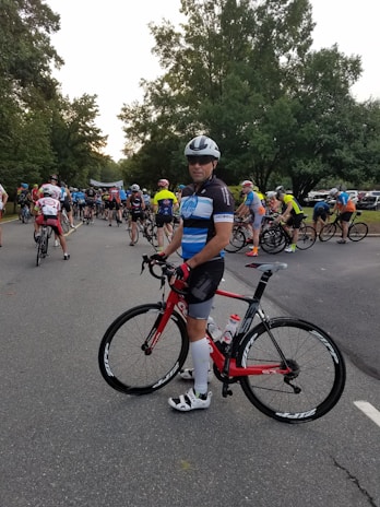 A group of cyclists gathered on a road, with a prominent cyclist in the foreground holding a red bicycle. The participants wear colorful cycling attire and helmets, preparing for what seems to be a race or an organized ride event. Tall trees line the road, creating a lush green backdrop.