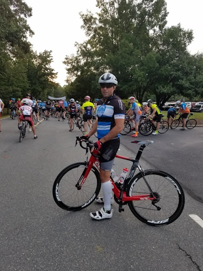 A group of cyclists gathered on a road, with a prominent cyclist in the foreground holding a red bicycle. The participants wear colorful cycling attire and helmets, preparing for what seems to be a race or an organized ride event. Tall trees line the road, creating a lush green backdrop.