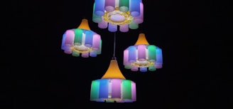 A set of colorful chandeliers with cylindrical lampshades in shades of pink, green, and blue, hanging against a dark background. The design features yellow accents and a warm central light source.
