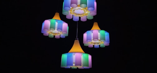 A set of colorful chandeliers with cylindrical lampshades in shades of pink, green, and blue, hanging against a dark background. The design features yellow accents and a warm central light source.